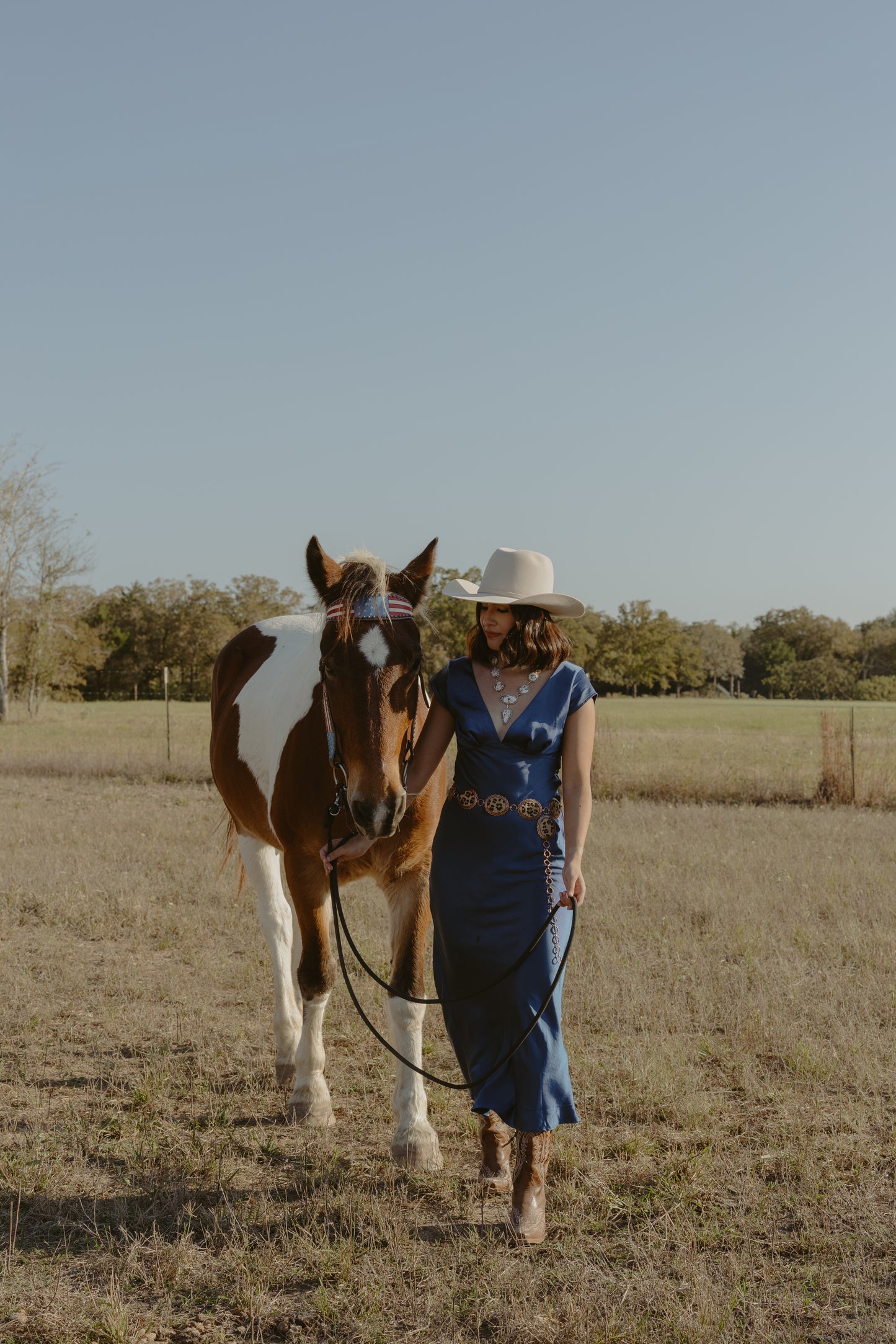 Woman in a blue dress and white hat walking with a brown and white horse in a field.